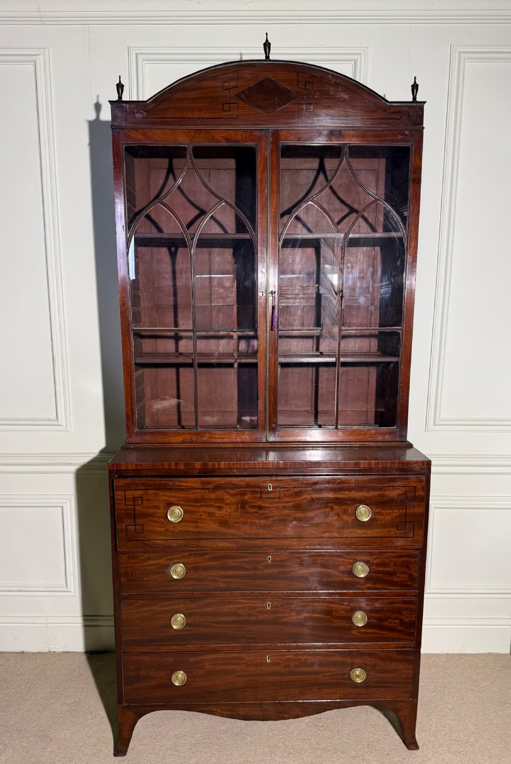 A tall Georgian mahogany secretaire bookcase featuring upper glazed doors with gothic arch bars, an arched pediment with finials, and a lower chest of drawers with brass ring handles.