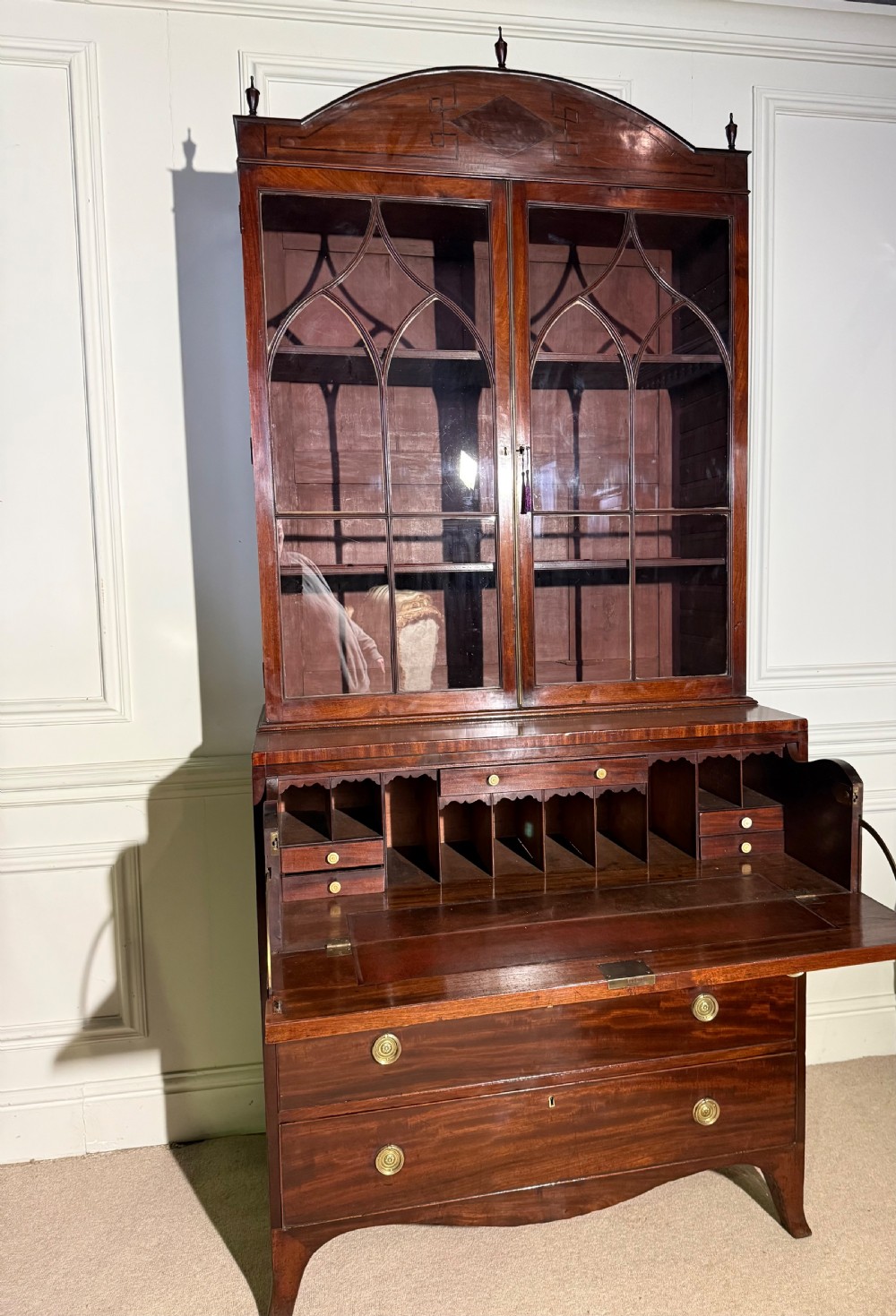 A tall Georgian mahogany secretaire bookcase featuring upper glazed doors with gothic arch bars, an arched pediment with finials, and a lower chest of drawers with brass ring handles. View IX