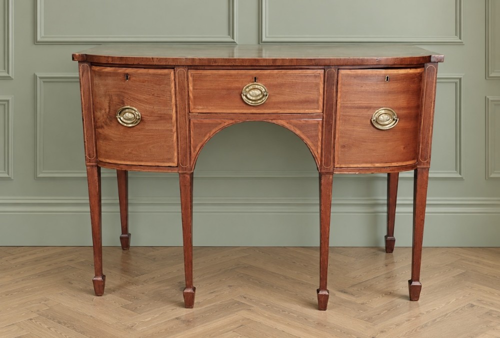 An 18th-century Sheraton style mahogany sideboard featuring brass oval drop handles, boxwood stringing, a central arch drawer, and square tapered legs ending in spade feet.