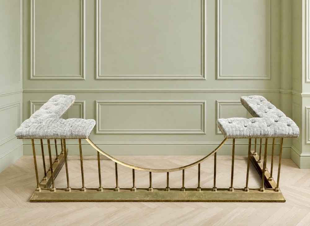A solid brass club fender featuring a dipped central rail, vertical spindle supports, and button-tufted light grey upholstered corner seats.
