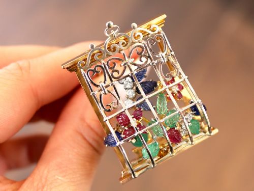 fig 1. An 18ct gold window-framed brooch featuring carved emerald, ruby, and sapphire floral elements behind a silver-coloured metal grate.