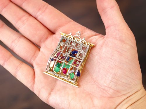 fig 2. An 18ct gold window-framed brooch featuring carved emerald, ruby, and sapphire floral elements behind a silver-coloured metal grate.
