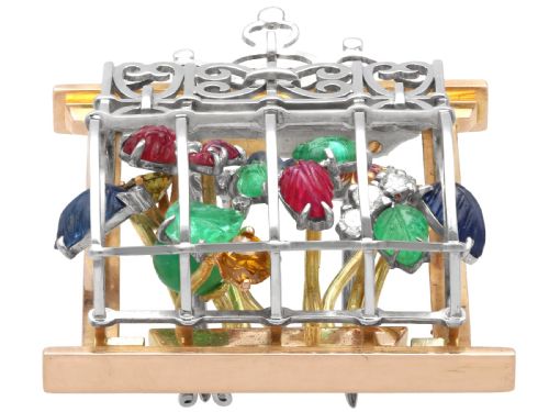 fig 3. An 18ct gold window-framed brooch featuring carved emerald, ruby, and sapphire floral elements behind a silver-coloured metal grate.