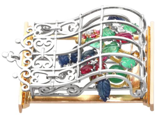 fig 4. An 18ct gold window-framed brooch featuring carved emerald, ruby, and sapphire floral elements behind a silver-coloured metal grate.