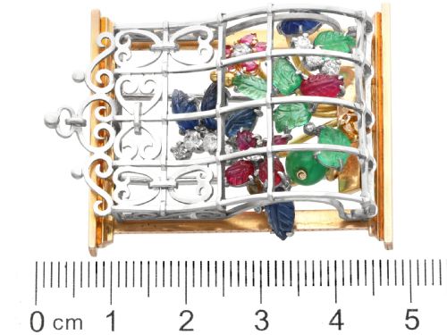 fig 8. An 18ct gold window-framed brooch featuring carved emerald, ruby, and sapphire floral elements behind a silver-coloured metal grate.