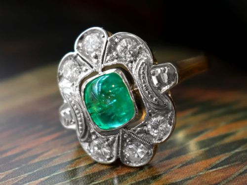 fig 2. An Art Deco dress ring featuring a central cushion-cut green emerald surrounded by a geometric pierced diamond halo set in white metal on an 18ct yellow gold band.