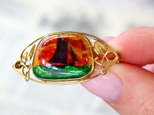 fig 3. An Arts and Crafts 15ct yellow gold brooch featuring a central enamel panel depicting a sailboat against a sunset sky framed by stylized gold leaves.