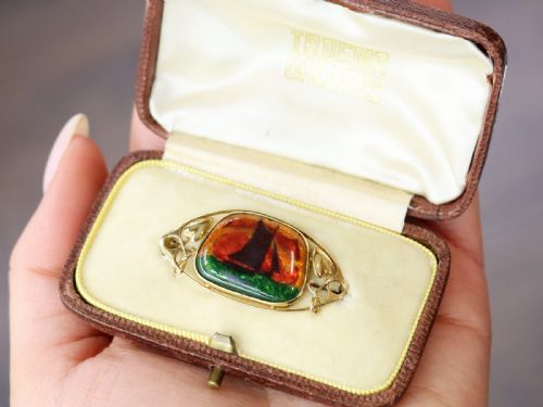 fig 4. An Arts and Crafts 15ct yellow gold brooch featuring a central enamel panel depicting a sailboat against a sunset sky framed by stylized gold leaves.