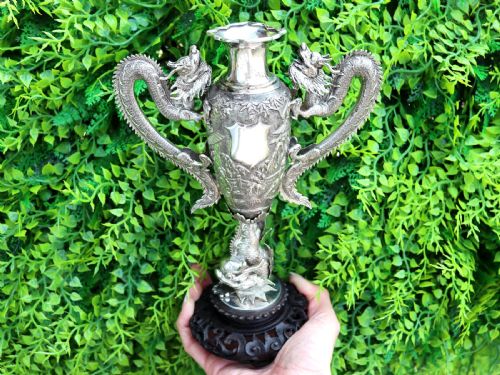 fig 1. A silver trophy-style vase featuring sculpted dragon handles, relief carving, and a dark wooden base.