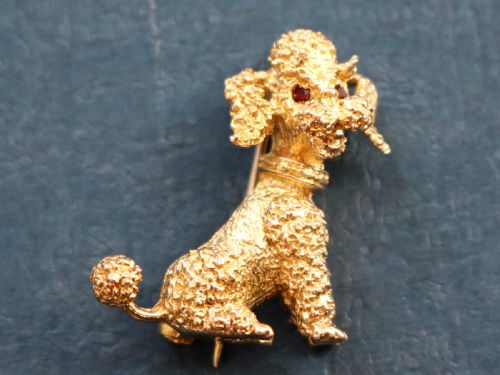 fig 2. A 9ct yellow gold brooch sculpted as a seated poodle with textured fur and red garnet eyes holding a stick in its mouth.