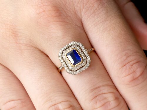 fig 2. An 18ct yellow gold ring featuring a central emerald-cut blue sapphire framed by a geometric double halo of round diamonds with millegrain edging.