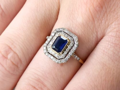 fig 3. An 18ct yellow gold ring featuring a central emerald-cut blue sapphire framed by a geometric double halo of round diamonds with millegrain edging.