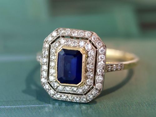 fig 4. An 18ct yellow gold ring featuring a central emerald-cut blue sapphire framed by a geometric double halo of round diamonds with millegrain edging.
