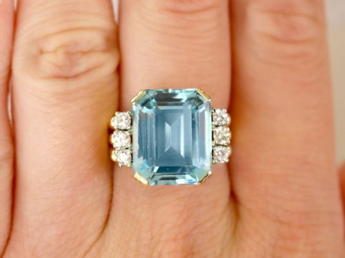 fig 2. A 14ct yellow gold dress ring featuring a large emerald-cut light blue aquamarine central gemstone flanked by six round diamonds.