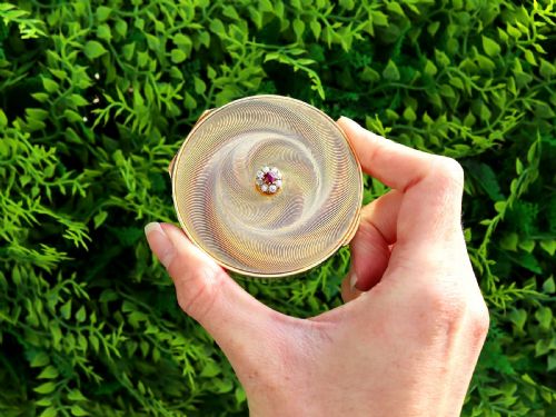 fig 2. A circular 9ct yellow gold powder compact featuring an engine-turned swirling pattern and a central ruby and diamond cluster.