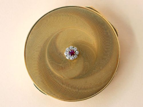 fig 5. A circular 9ct yellow gold powder compact featuring an engine-turned swirling pattern and a central ruby and diamond cluster.