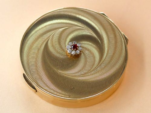 fig 6. A circular 9ct yellow gold powder compact featuring an engine-turned swirling pattern and a central ruby and diamond cluster.
