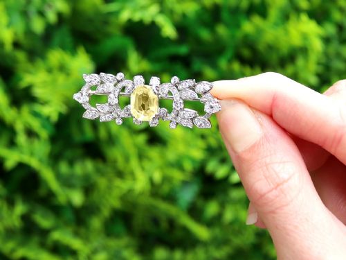fig 1. An Art Deco platinum brooch centered with an oval cushion-cut yellow sapphire enclosed by diamond-set scrollwork and foliate motifs.