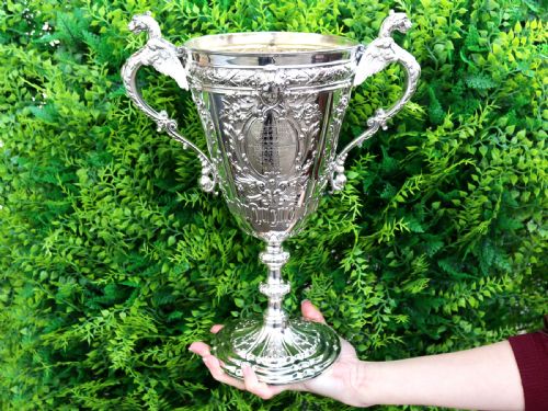 fig 1. An antique Victorian sterling silver presentation cup featuring ornate chased scrollwork, twin handles topped with winged figures, and a stepped pedestal base.
