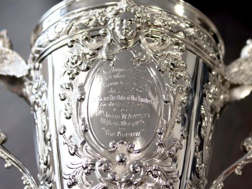 fig 2. An antique Victorian sterling silver presentation cup featuring ornate chased scrollwork, twin handles topped with winged figures, and a stepped pedestal base.