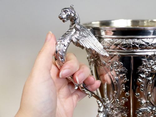 fig 3. An antique Victorian sterling silver presentation cup featuring ornate chased scrollwork, twin handles topped with winged figures, and a stepped pedestal base.