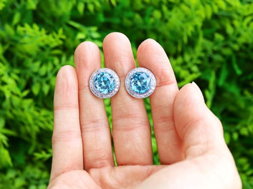 fig 1. A pair of 14ct white gold stud earrings featuring round blue zircon gemstones encircled by diamond halos with millegrain borders.