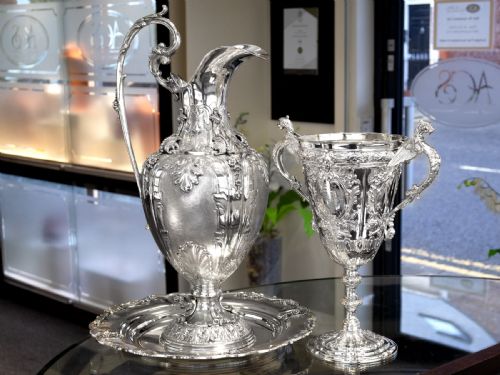 fig 4. An antique Victorian sterling silver presentation cup featuring ornate chased scrollwork, twin handles topped with winged figures, and a stepped pedestal base.