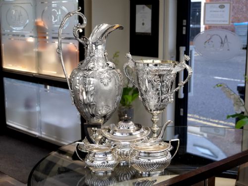 fig 5. An antique Victorian sterling silver presentation cup featuring ornate chased scrollwork, twin handles topped with winged figures, and a stepped pedestal base.