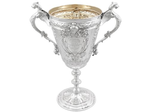 fig 6. An antique Victorian sterling silver presentation cup featuring ornate chased scrollwork, twin handles topped with winged figures, and a stepped pedestal base.