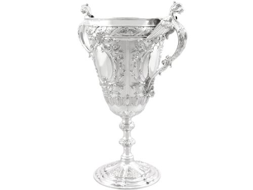 fig 7. An antique Victorian sterling silver presentation cup featuring ornate chased scrollwork, twin handles topped with winged figures, and a stepped pedestal base.