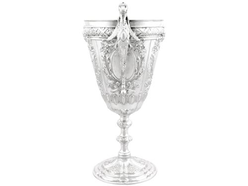 fig 8. An antique Victorian sterling silver presentation cup featuring ornate chased scrollwork, twin handles topped with winged figures, and a stepped pedestal base.