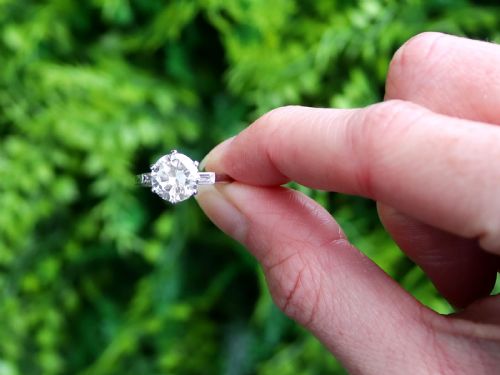 fig 1. A platinum solitaire ring featuring a large round cut central diamond set with six claws, flanked by step-cut baguette diamonds on each shoulder.