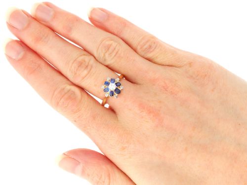 fig 8. An 18ct yellow gold ring featuring a central round diamond surrounded by six oval blue sapphires and outer round diamond accents arranged in a flower motif.