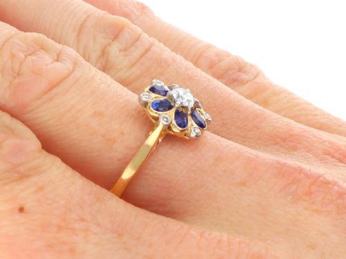 fig 9. An 18ct yellow gold ring featuring a central round diamond surrounded by six oval blue sapphires and outer round diamond accents arranged in a flower motif.