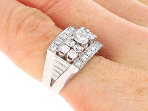 fig 8. An Art Deco platinum dress ring featuring a central round diamond bordered by multiple smaller old-cut diamonds in a geometric rectangular mount with millegrain edges.