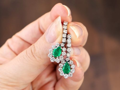fig 3. A pair of 18ct white gold drop earrings featuring pear-cut green emeralds surrounded by round diamond halos suspended from vertical diamond bars.