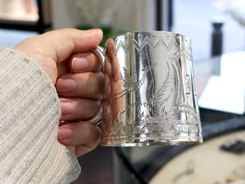 fig 2. A tapered sterling silver christening mug featuring hand-engraved foliage and bird motifs, geometric borders, a loop handle, and a gilt interior.