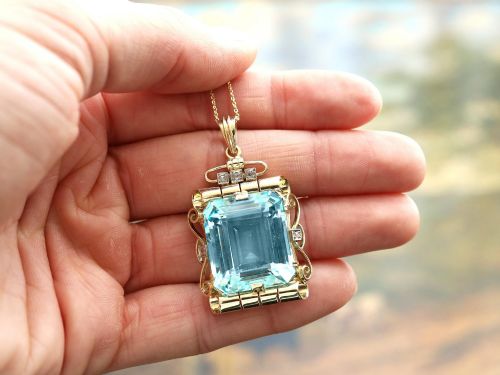 fig 2. A large emerald-cut aquamarine gemstone mounted in a yellow gold scrollwork frame accented with small diamonds and suspended from a chain.