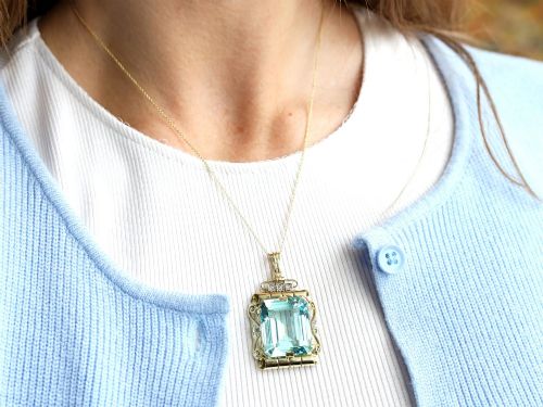 fig 4. A large emerald-cut aquamarine gemstone mounted in a yellow gold scrollwork frame accented with small diamonds and suspended from a chain.