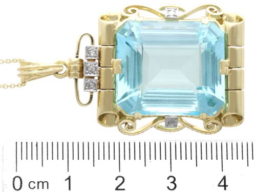 fig 9. A large emerald-cut aquamarine gemstone mounted in a yellow gold scrollwork frame accented with small diamonds and suspended from a chain.