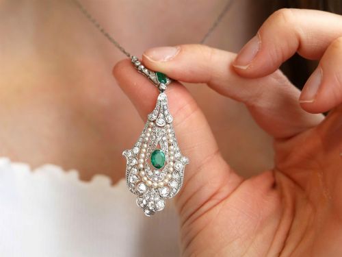 fig 4. An openwork platinum pendant featuring an elongated drop shape set with diamonds, a line of seed pearls, and two vibrant green Colombian emeralds.