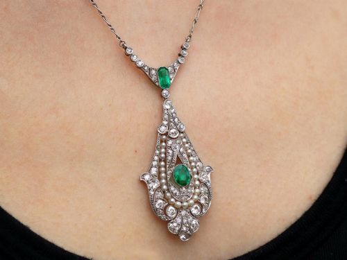 fig 11. An openwork platinum pendant featuring an elongated drop shape set with diamonds, a line of seed pearls, and two vibrant green Colombian emeralds.