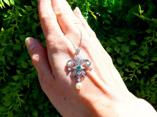 fig 1. An Art Deco platinum pendant shaped as a stylised fleur-de-lis, embellished with pave-set diamonds, a central rectangular emerald, and a hanging pearl drop.