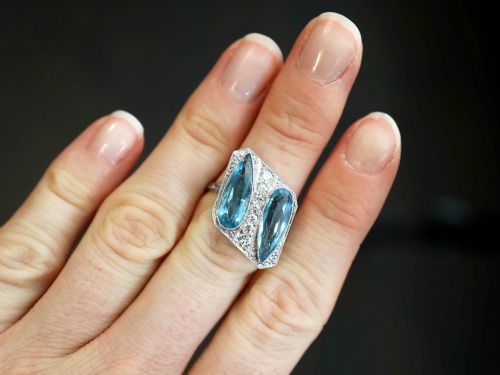 fig 2. An Art Deco platinum cocktail ring featuring two pear-cut light blue aquamarines set diagonally beside a central row of round diamonds with millegrain borders.