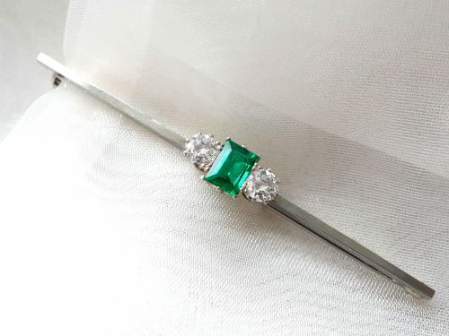 fig 4. An antique 18ct white gold bar brooch featuring a central rectangular emerald cut emerald flanked by two round diamonds on a slender horizontal metal bar.