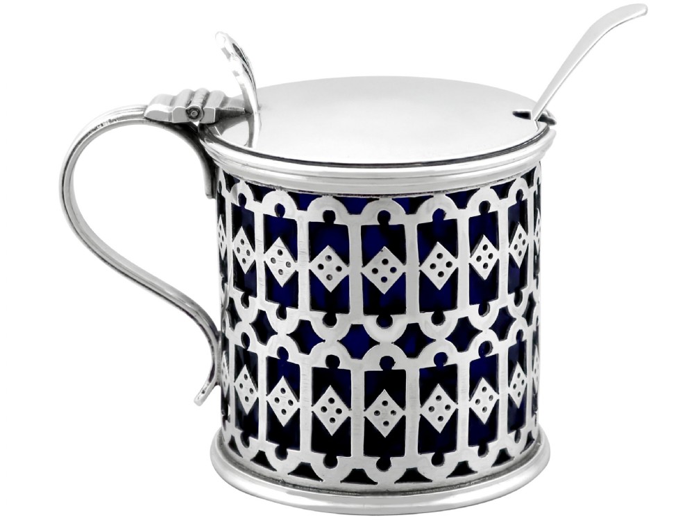 A pierced sterling silver mustard pot featuring geometric openwork, a cobalt blue glass liner, a flat hinged lid, and an accompanying spoon.