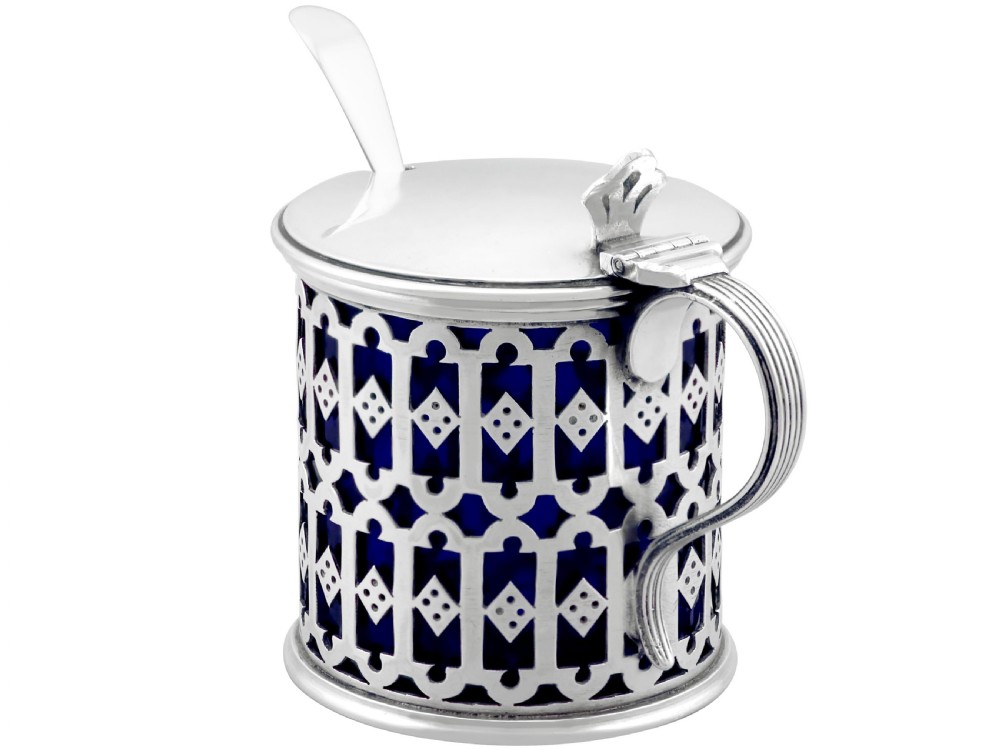 A pierced sterling silver mustard pot featuring geometric openwork, a cobalt blue glass liner, a flat hinged lid, and an accompanying spoon. View VI