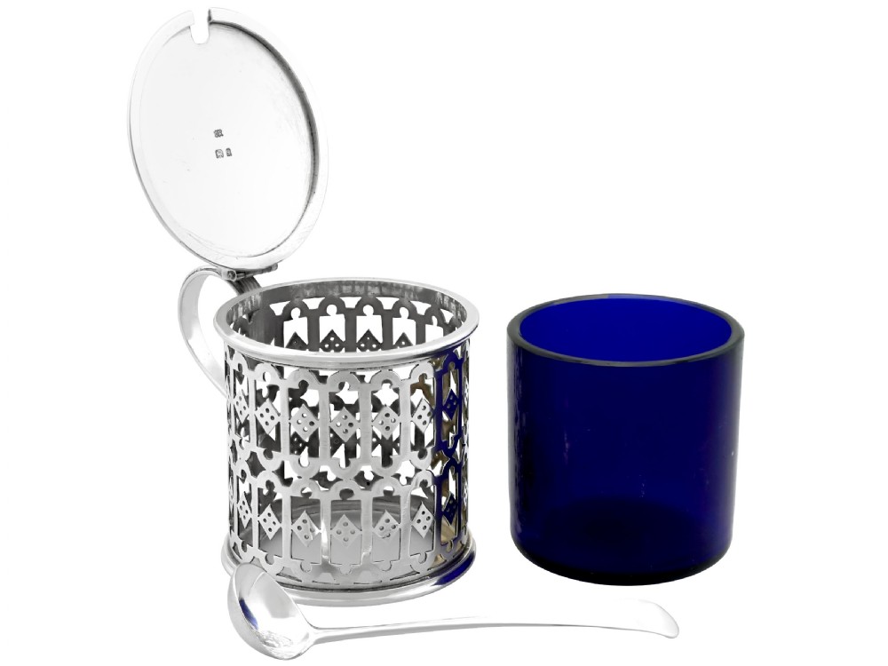 A pierced sterling silver mustard pot featuring geometric openwork, a cobalt blue glass liner, a flat hinged lid, and an accompanying spoon. View VII