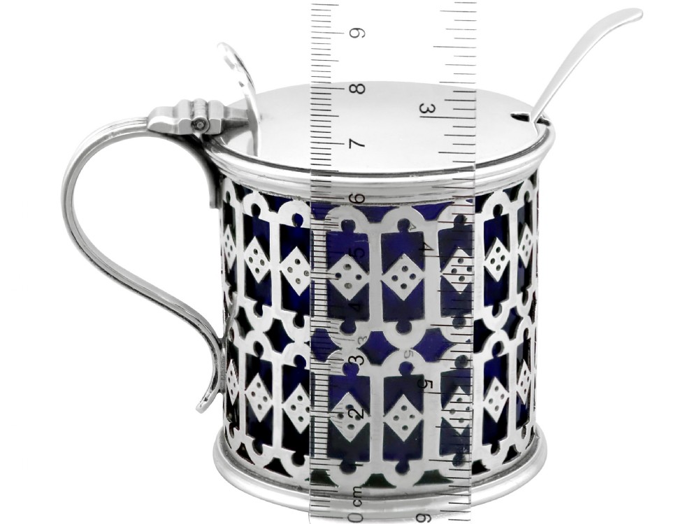 A pierced sterling silver mustard pot featuring geometric openwork, a cobalt blue glass liner, a flat hinged lid, and an accompanying spoon. View VIII