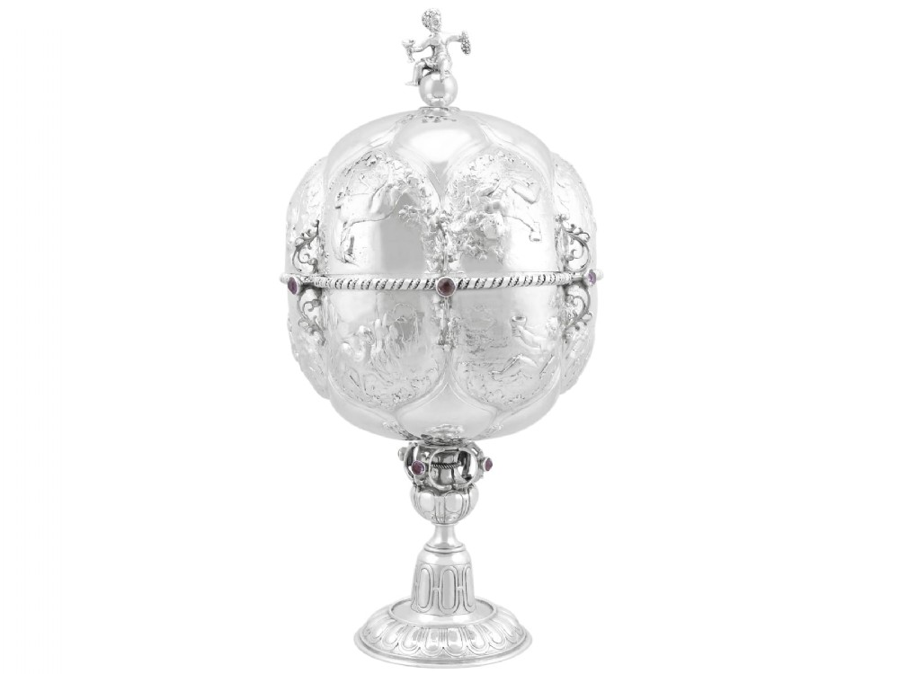 An antique German sterling silver covered presentation cup featuring chased relief panels, set cabochon gemstones, and a putto finial.